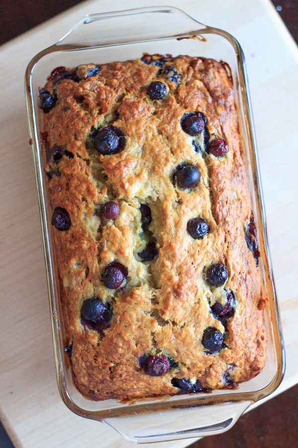 Blueberry banana bread baked in clear bread dish sitting on wood cutting board