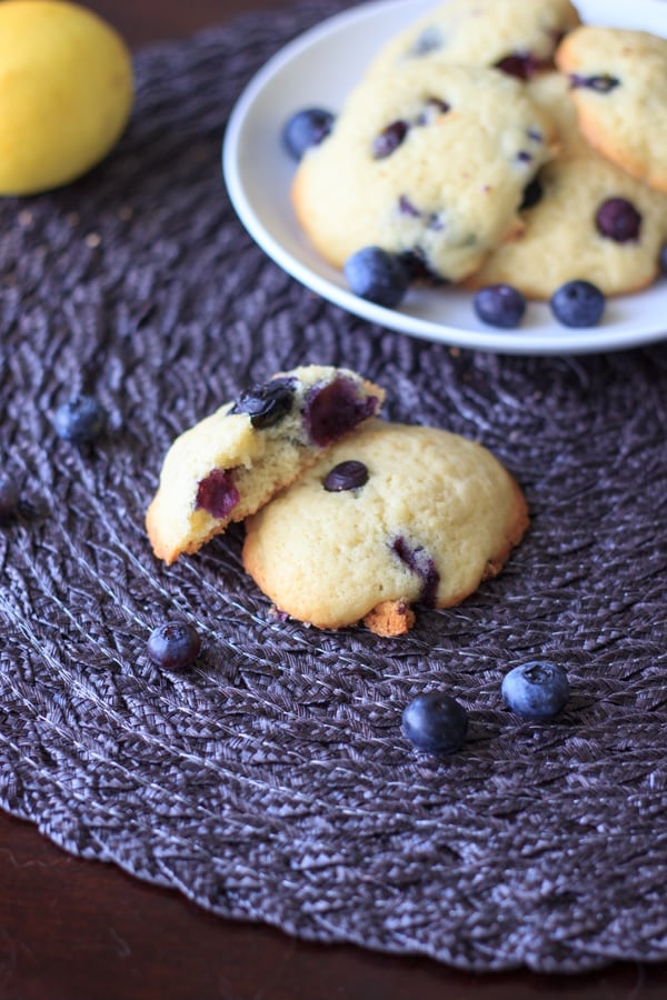 Lemon blueberry cookie in half, showing inside texture, with plate of cookies and lemon in background 