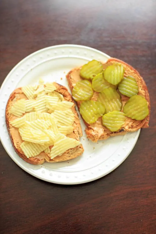 Peanut butter on two slices of bread, sliced pickles on one side and potato chips on the other, open-faced sandwich on white plate