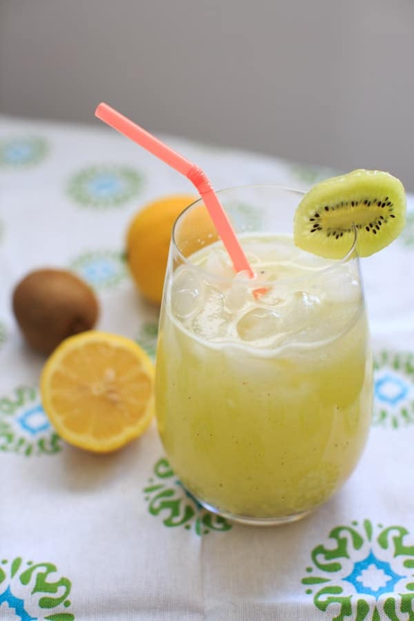 Homemade Kiwi Lemonade with slice of kiwi on edge of clear drinking glass, half a lemon and kiwi in background