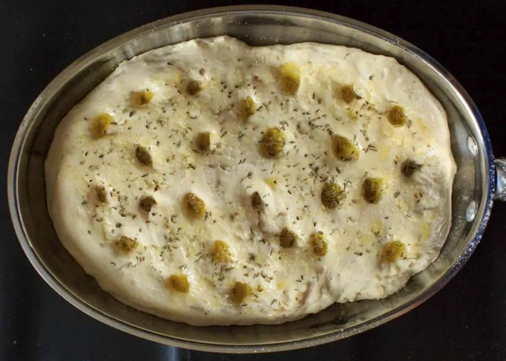 garlic herb focaccia bread dough in baking dish with holes poked in and olive oil sprinkked on top