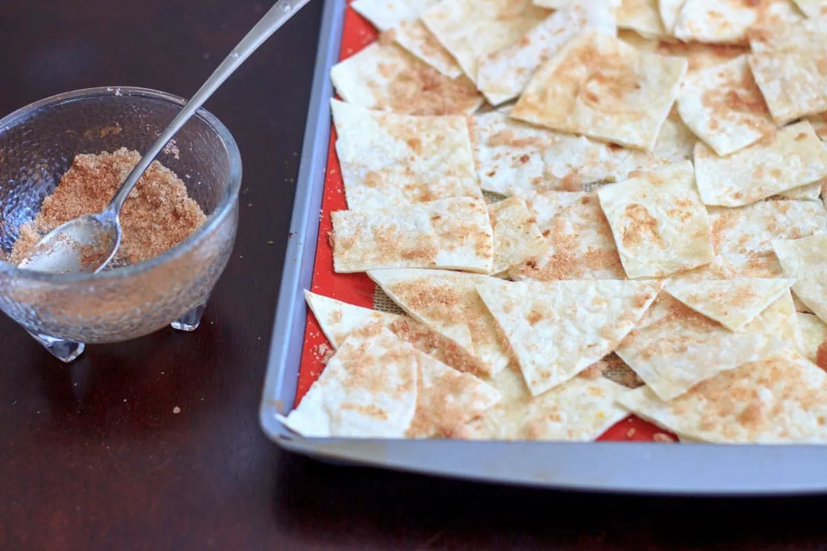 cut up tortillas on a baking pan with bowl of cinnamon sugar and spoon next to it