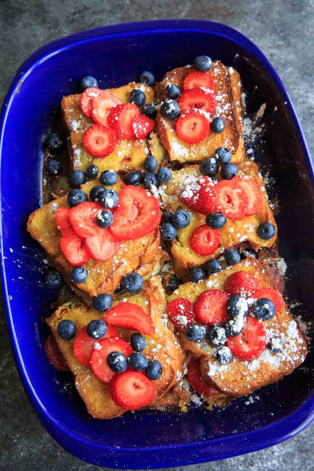 blue casserole dish filled with stuffed french toast, topped with blueberries, strawberries and powdered sugar