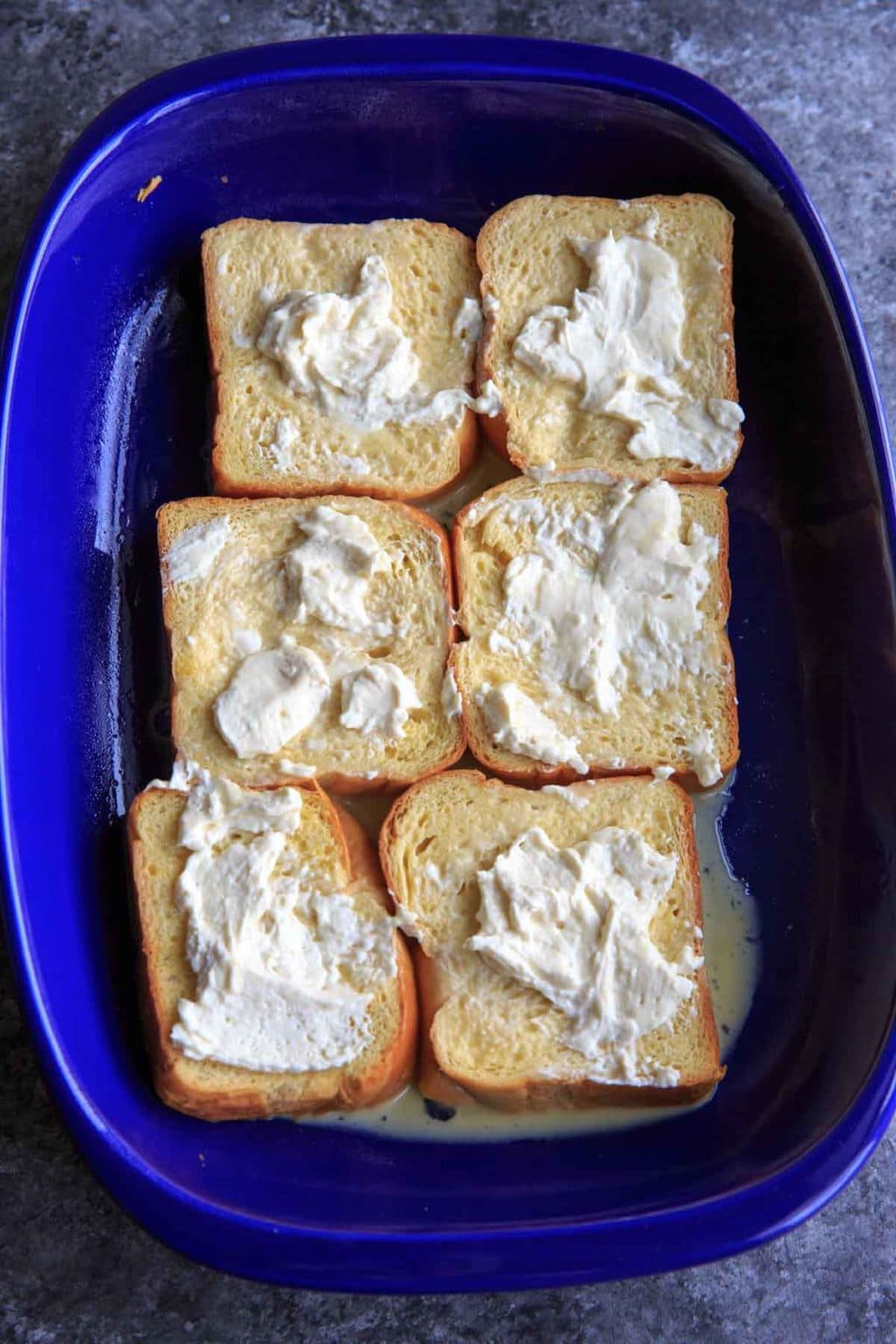 pieces of bread for french toast topped with cream cheese and egg in blue casserole dish
