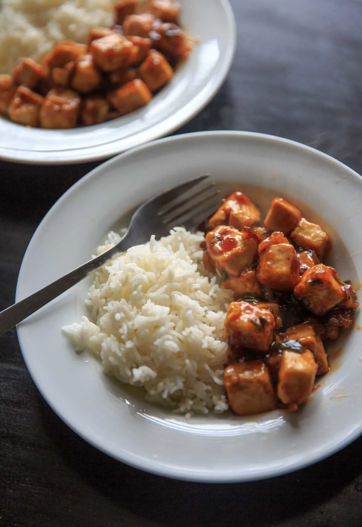 General Tso's Tofu serve with rice in white bowl with fork and second plate serving in background