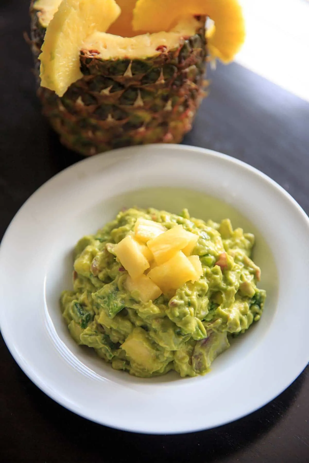 Pineapple Jalapeno Guacamole on white plate topped with pineapple chunks and view of whole pineapple in background