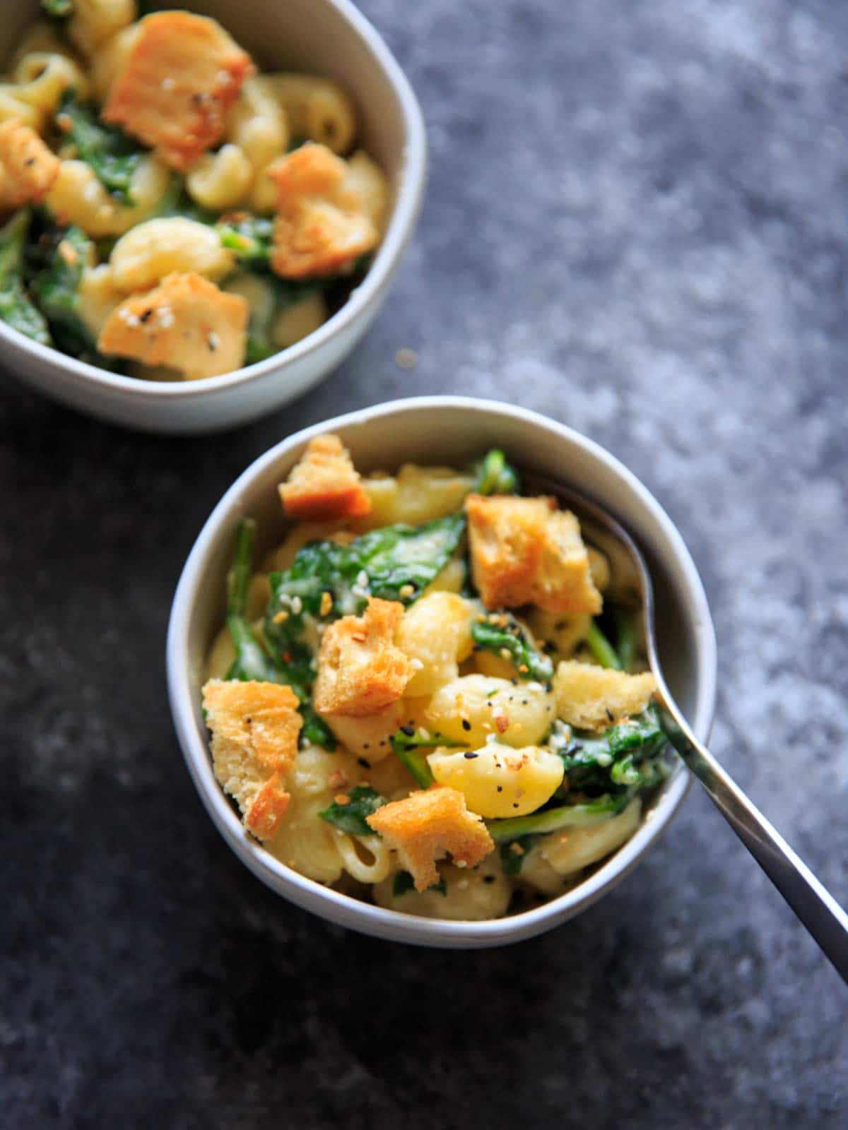 White cheddar macaroni and cheese with spinach and topped with "everything bagel" seasoned homemade croutons, in two white small bowls one with a fork in it