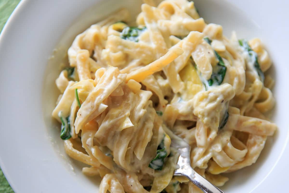 landscape view picture of cooked pasta in white bowl with spinach artichoke sauce, with fork twirled in pasta