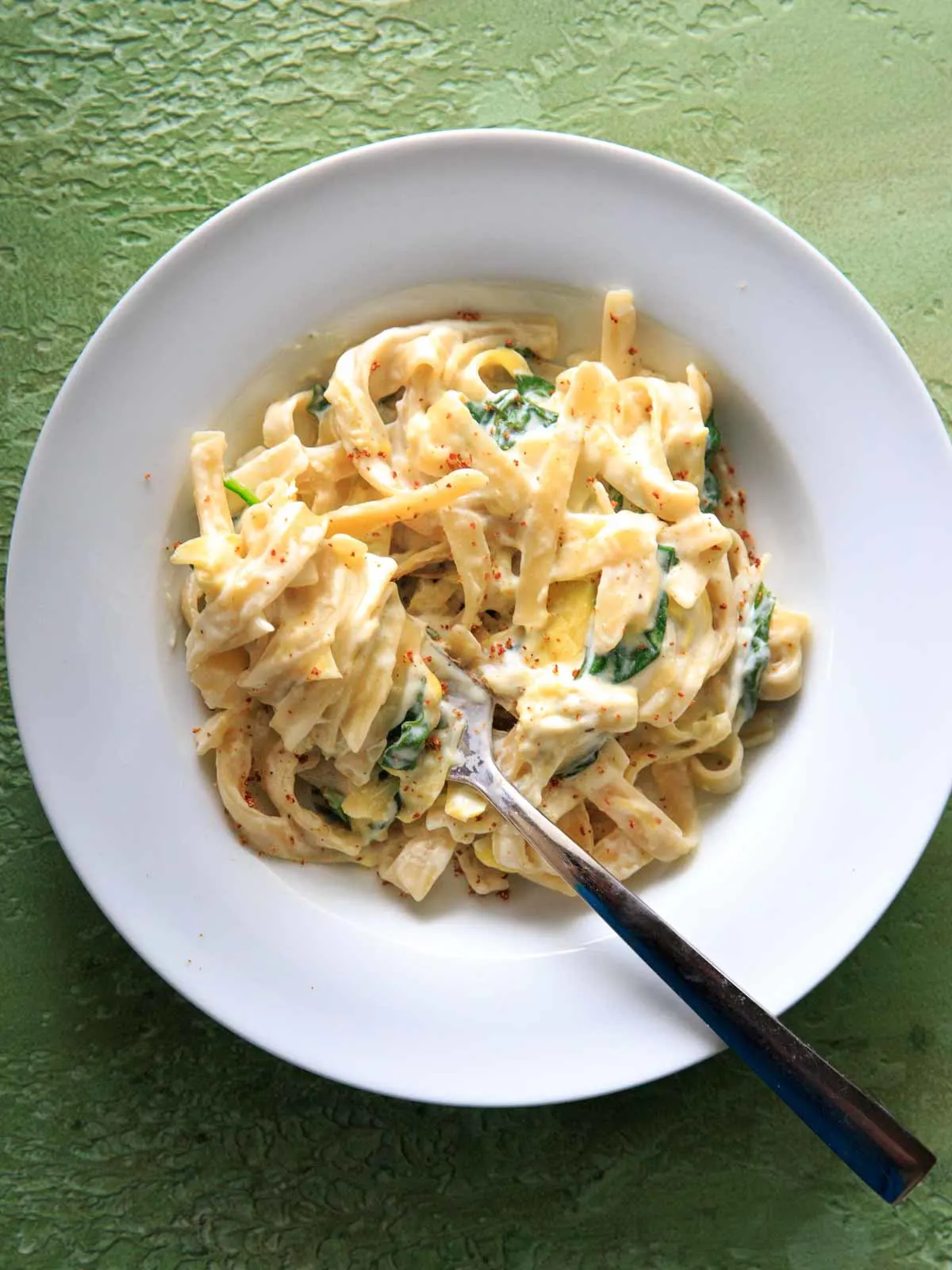 white plate filled with serving of spinach and artichoke pasta with fork twirled in, topped with sprinkled red pepper flakes