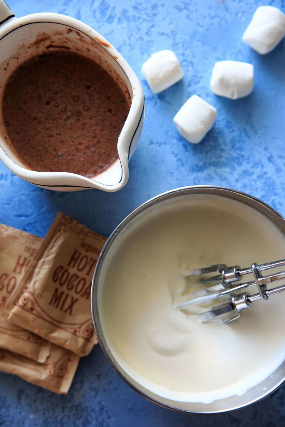 hot cocoa mix packets laying next to a mixing bowl of whipped whipping cream and sweetened condensed milk, and a pour measuring cup of hot cocoa