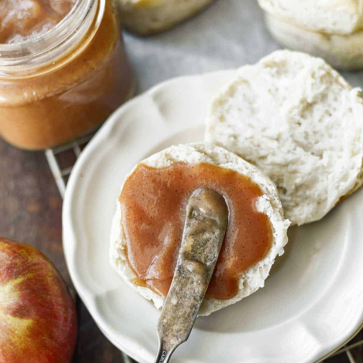 knife spreading apple butter on half a roll on white plate.